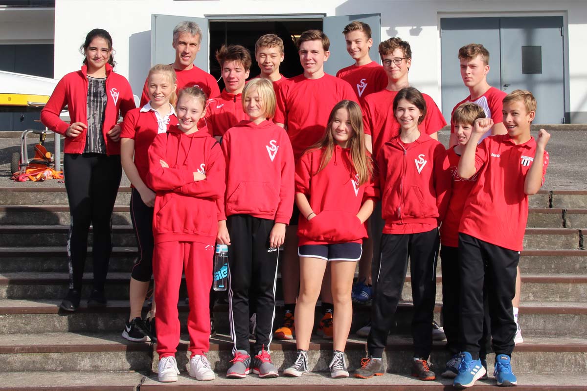 Jugend-Rudermannschaft mit Trainer in roten Outfits vor dem Bootshaus.