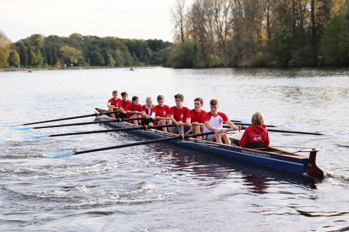 Achter-Ruderboot mit Jugendlichen in Rot auf ruhigem Fluss.